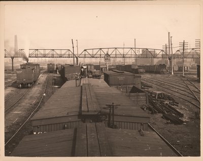 Horizontal, black and white photograph of rail cars in a rail yard. A man sits on top of one of the cars by William G. Swekosky